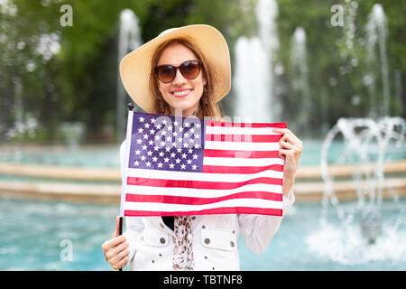 Happy young woman in sunglasses holding USA flag in park at fountains Stock Photo