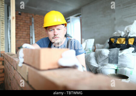 Portrait of bearded builder wearing yellow hardhat and protective gloves laying red blocks on concrete wall. Smart male trying to put as strict as pos Stock Photo