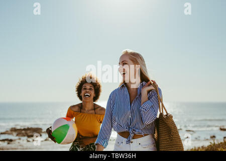 Cheerful young women walking with a ball and bag along the sea shore. Female friends on beach vacation.