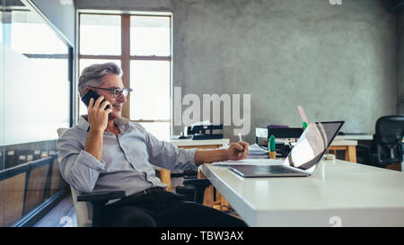 businessman on the phone sitting at the laptop in his office. Male business professional in office talking on cell phone. Stock Photo
