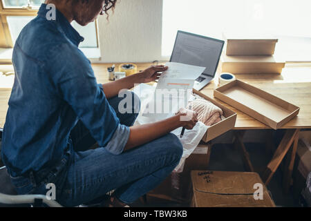 Female entrepreneur reading and verifying the invoice before shipping the product to the customer. Woman preparing shipment for delivery at her desk. Stock Photo