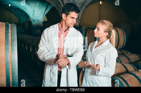 Friendly smiling positive specialists in white robes checking ageing process of wine Stock Photo