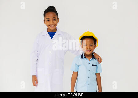 African American girl in scientist uniform and little boy in engineer hardhat smiling and looking at camera against white background Stock Photo