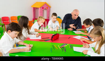 Friendly male teacher talking to children, sitting together around desk in classroom Stock Photo