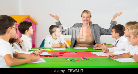 Friendly female teacher talking to children, sitting together around desk in the classroom Stock Photo