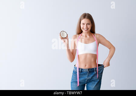 Beautiful young woman in loose jeans, with coconut and measuring tape on light background. Weight loss concept Stock Photo