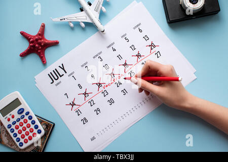 Hand of woman planning her vacation on calendar. Travel preparation: camera and passport on blue background. Flat lay. Top view. Stock Photo