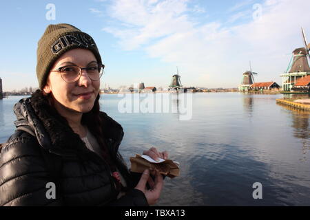 Caucasian tourist girl dressed in winter clothes walks among the canals of the Zaan river, in Zaansche Schans, among the old Dutch windmills Stock Photo