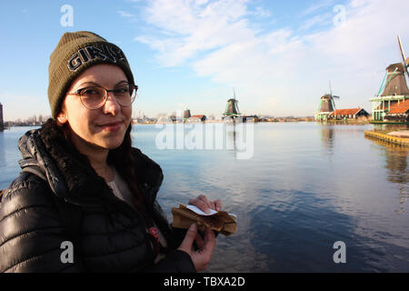 Caucasian tourist girl dressed in winter clothes walks among the canals of the Zaan river, in Zaansche Schans, among the old Dutch windmills Stock Photo