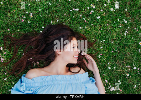 Beautiful young brunette girl in blue dress looking away and lying on a clearing among white flowers
