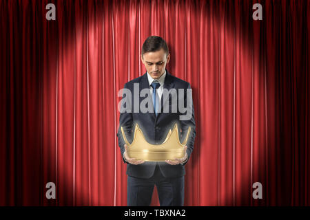 Knee-deep front view of businessman looking down at gold crown which he is holding in hands, at red stage curtain. Stock Photo
