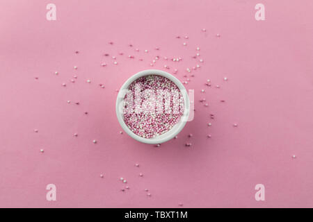 Sprinkles isolated on pink background - little white bowl with pearly pink cake topping sprinkles and some scattered on pink - top view Stock Photo