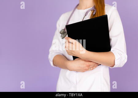 Hands of young female doctor in white uniform with phonendoscope on her neck holding black folder on purple background with space for text. Healthcare Stock Photo