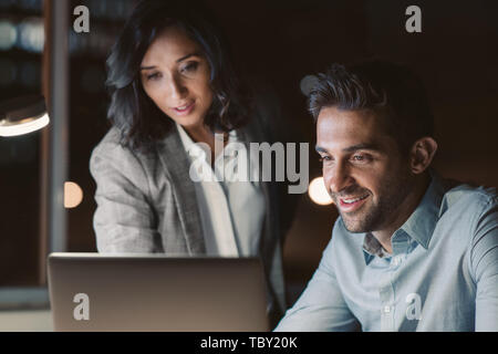 Smiling office colleagues using a laptop while working overtime together Stock Photo