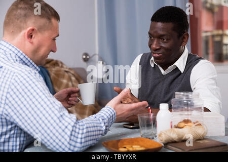 Interracial two male friends drinking coffee and talking in home interior Stock Photo
