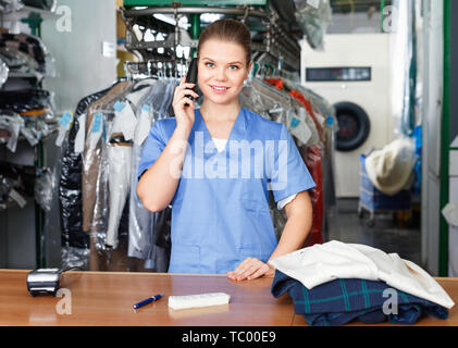 Smiling diligent cheerful  girl worker of laundry talking on phone while checking clothes taken for dry cleaning Stock Photo