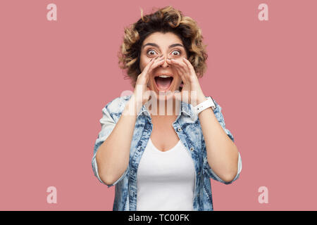 Portrait of shocked young woman with curly hairstyle in casual blue shirt standing, screaming, sharing something and looking at camera with amazed fac Stock Photo