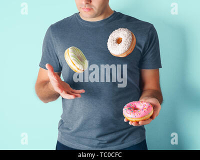 Juggler man juggles donuts on blue background. Doughnuts delivery or unhealthy diet concept. Flying glazed donuts with sprinkles and desiccated coconut in male hands Stock Photo