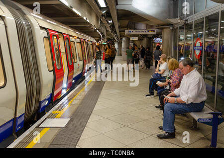 Passengers sit and wait on a bench on the platform at Westminster London Underground Station as other passengers board a train. Stock Photo