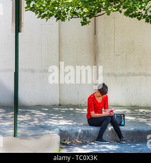 ASHEVILLE, NC, USA-5/31/19: A solitary young woman in red shirt sits on city street curb looking at smart phone, framed by tree limb and street sign.