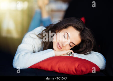 An attractive smiling woman lies on grey couch with pillows at cozy home in the morning. Cute girl having rest at home in lazy day. Stock Photo