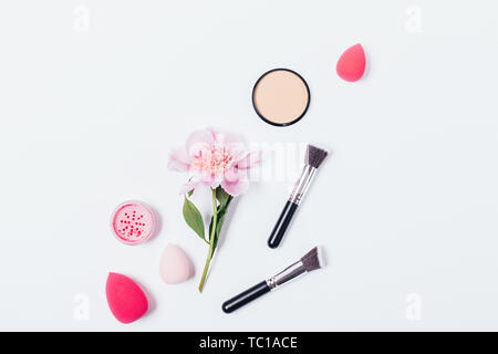 Flat lay abstract makeup background of pink peony, cosmetic brushes and sponges next to powder and blush on white background. Stock Photo