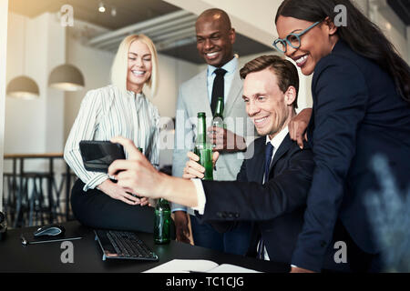Smiling businessman sitting at his desk talking a selfie with colleagues behind him in an office during after work drinks Stock Photo