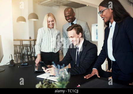 Laughing group of diverse businesspeople watching an office colleague playing a cellphone game while sitting at his desk after work Stock Photo