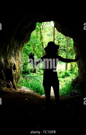 Slim girl silhouette at the entrance to natural cave in the forrest, young explorer Stock Photo