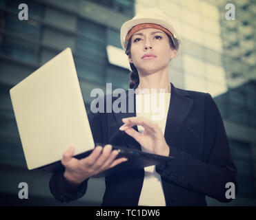 Smiling woman architector in suit and hat is exploring project in her laptop near the building. Stock Photo