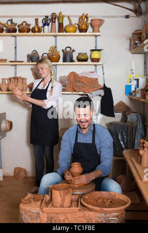 Smiling craftsman making pot using pottery wheel and various clay vessels Stock Photo