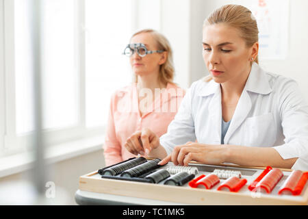 Eye measuring device. Focused woman observing her lens collection in wide case while patient sitting with glasses on Stock Photo