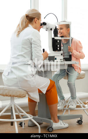 Scary apparatus. Beaming pretty child being in great mood while having session with focused professional doctor Stock Photo