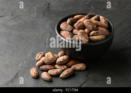 Roasted unpeeled cocoa beans in a black ceramic bowl next to a pile of unpeeled cocoa beans isolated on black slate. Stock Photo