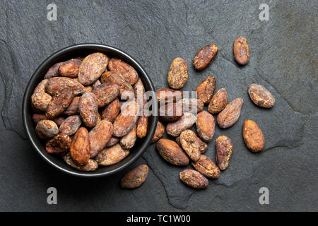 Roasted unpeeled cocoa beans in a black ceramic bowl next to unpeeled cocoa beans isolated on black slate from above. Stock Photo