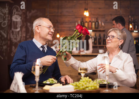 Woman in her sixties happy to recieve roses from her husband during dinner. Senior couple date. Fresh grapes. Cheerful old couple. Stock Photo