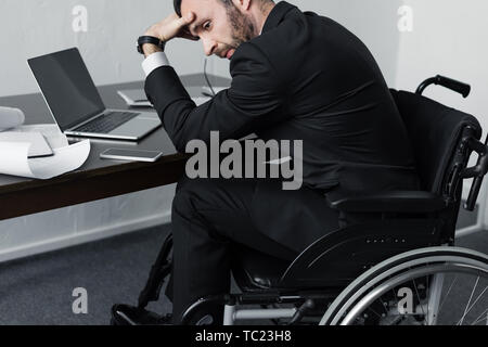 upset disabled businessman sitting at workplace in wheelchair and holding hand on forehead Stock Photo