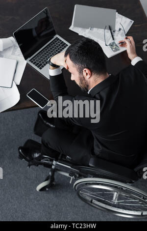 overhead view of disabled businessman sitting at workplace near papers, laptop and smartphone with blank screen