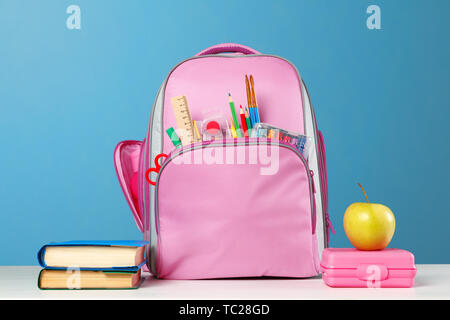 Student set. Pink backpack with stationery, a stack of books, a lunchbox, an apple on the table on a blue background. Back to school. Stock Photo