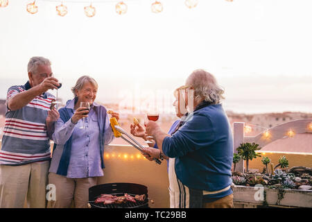 Happy senior friends having doing barbecue party on patio - Retired people having fun drinking wine and grilling meat at bbq dinner at home Stock Photo