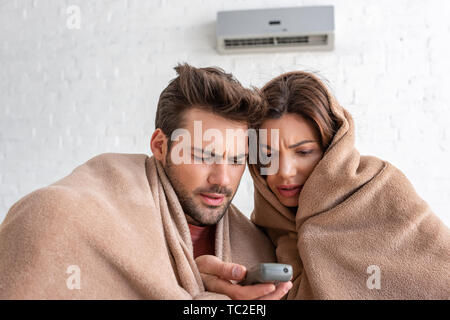 frozen man and woman warming under blankets while sitting under air conditioner with remote control Stock Photo