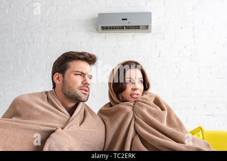 frozen man and woman warming under blankets while sitting under air conditioner Stock Photo
