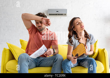 pretty woman with hand fan and handsome man with newspaper suffering from heat at home Stock Photo
