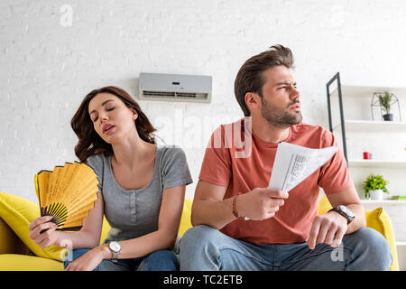 young woman with hand fan and handsome man with newspaper suffering from heat at home Stock Photo