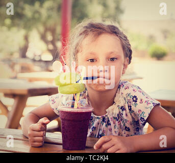 Fun cute thinking kid girl drinking smoothie juice with serious look in summer cafe. Closeup toned vintage portrait Stock Photo