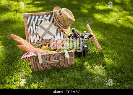 Vintage style summer vegetarian picnic in a wicker basket with old straw hat and cooler of beer bottles to the side on lush green grass outdoors Stock Photo