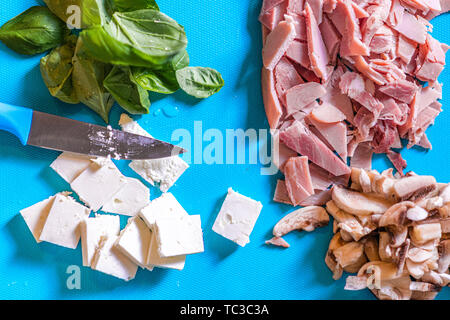 Cut up ingredients such as ham, mushroom, cheese and basil on a blue plastic chopping board with a short sharp knife Stock Photo