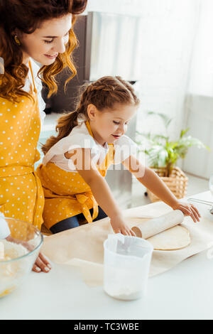 happy daughter in yellow polka dot apron rolling out dough on table next to mother Stock Photo