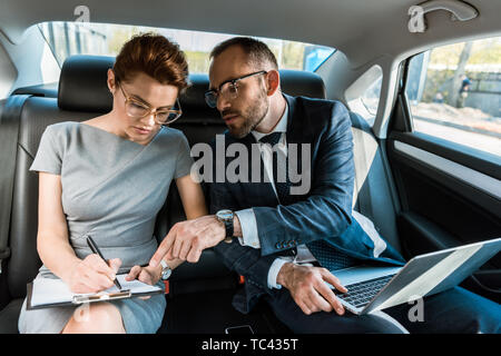 handsome businessman pointing with finger at clipboard while sitting with laptop near woman in car Stock Photo