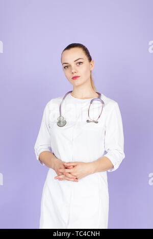 Young female blonde doctor with crossed hands with stethoscope on her neck on purple background with space for text. Doctod looks at camera Stock Photo